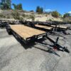Two black PJ flatbed trailers with wood decks and tandem axles parked on asphalt under blue sky