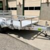 Aluminum utility trailer with open sides, black wheel, and rear ramp gate, parked on concrete near a building.
