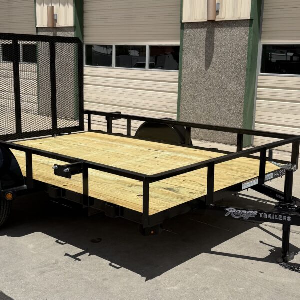 Utility trailer with a black frame and wood flooring sitting in the sun next to a closed multi-door garage at Pine Country.